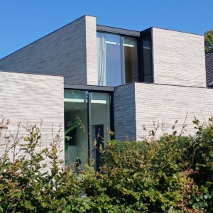 Nieuwbouwwoning in Lokeren uit rechthoekige blokken in lichtgrijze baksteen, met platte daken, achter een beukenhaag, een project van Bouwwerken Goossens