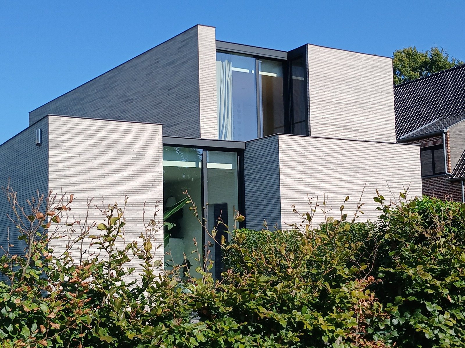 Nieuwbouwwoning in Lokeren uit rechthoekige blokken in lichtgrijze baksteen, met platte daken, achter een beukenhaag, een project van Bouwwerken Goossens
