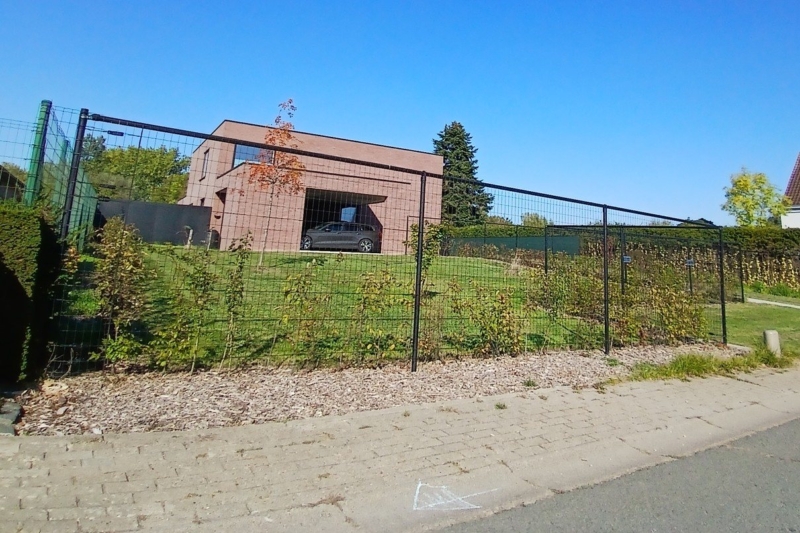Zijzicht op de nieuwbouwwoning in rode baksteen in Steenhuize, achter een draadafsluiting met jonge haag