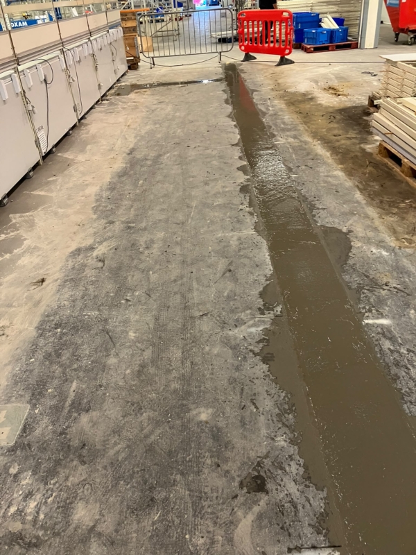 Afgezette werkzone in een bedrijfsruimte met een vers gegoten strook beton naast diepvriezers en rekken, een project van Bouwwerken Goossens