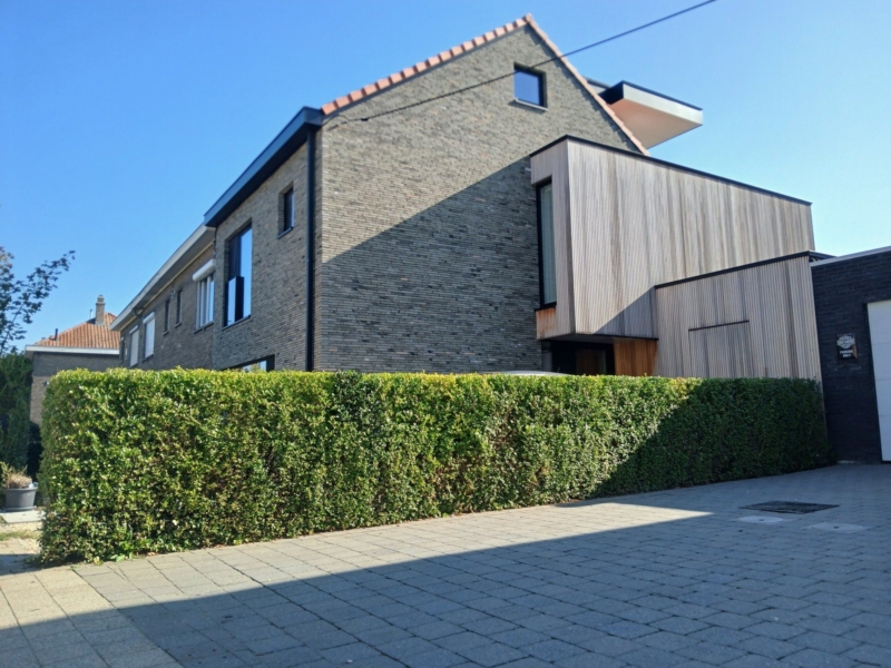 Verbouwde halfopen woning in Gentbrugge met een gevel in donkere baksteen, een uitbouw met verticale houten latten en een rood pannendak, een project van Bouwwerken Goossens